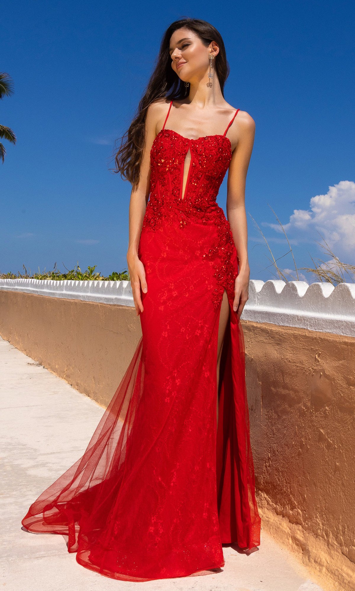 A woman stands outdoors under a sunny blue sky, wearing the Long Mermaid Prom Dress: Amelia Couture 7064—a red, sleeveless, floral lace gown with bead embellishments.